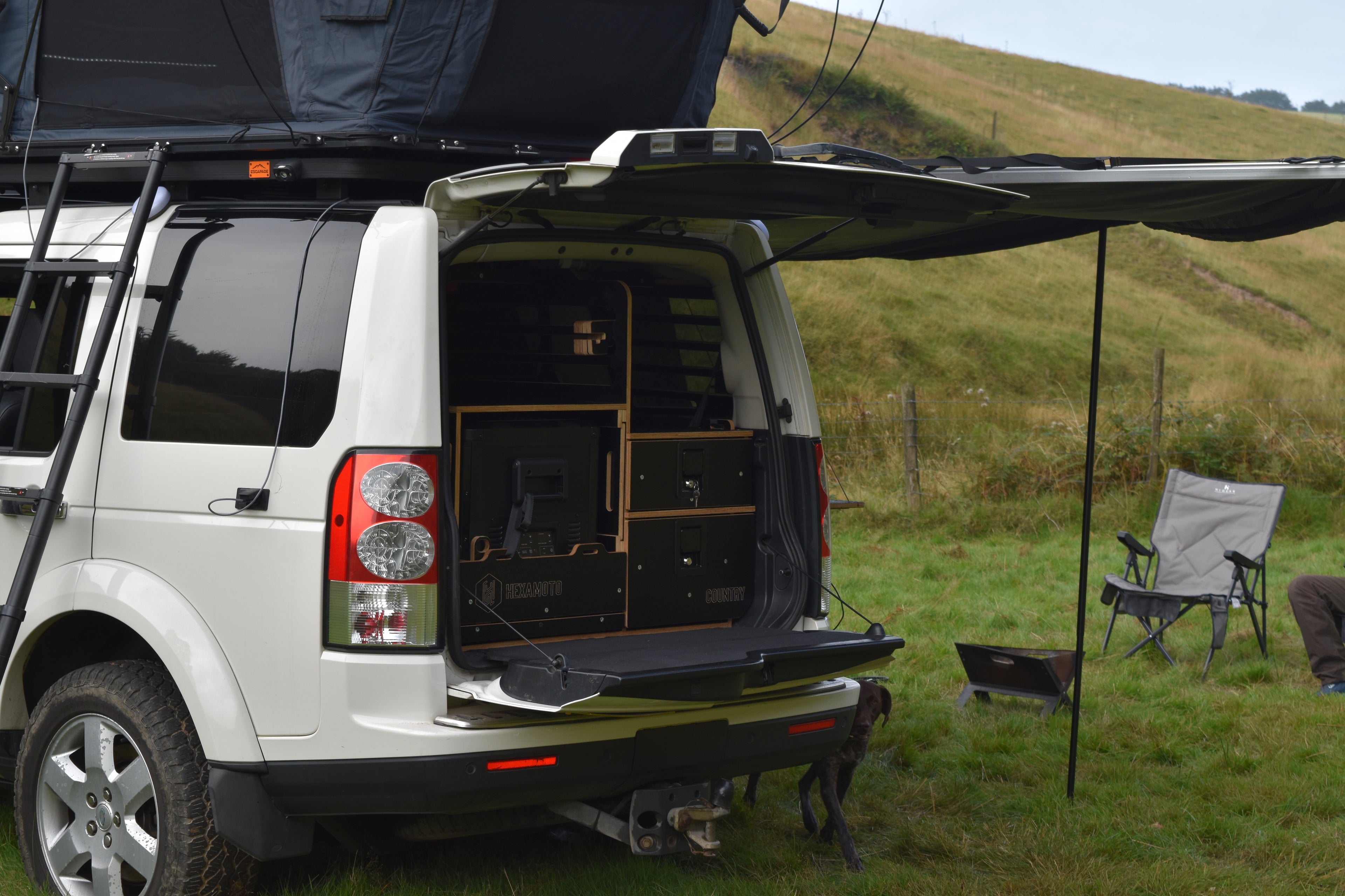 Discovery 4 with open rear doors, showing Hexamoto Country system and roof tent in a grassy outdoor setting