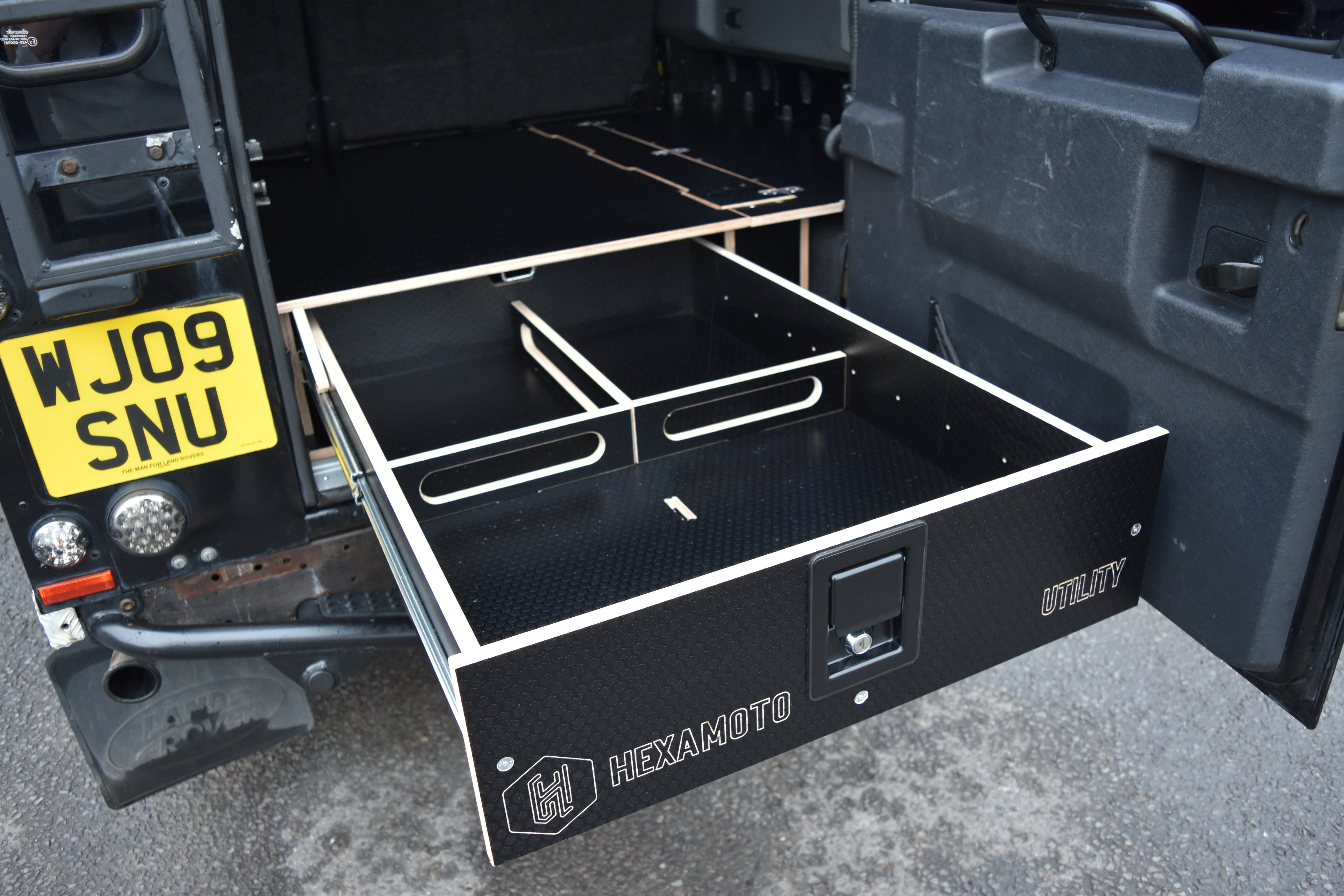 Black storage drawer with compartments attached to a vehicle, featuring the brand 'Hexamoto'.