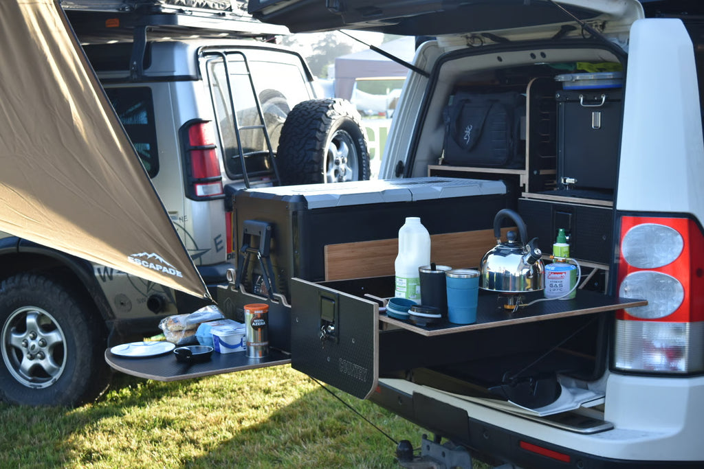 Trunk of a vehicle with camping gear and supplies, including a coolbox, bottles, and a kettle.