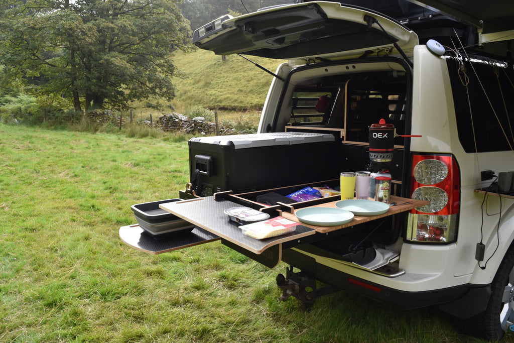 Car with an open tailgate displaying a Hexamoto Country camping setup in a grassy outdoor area.