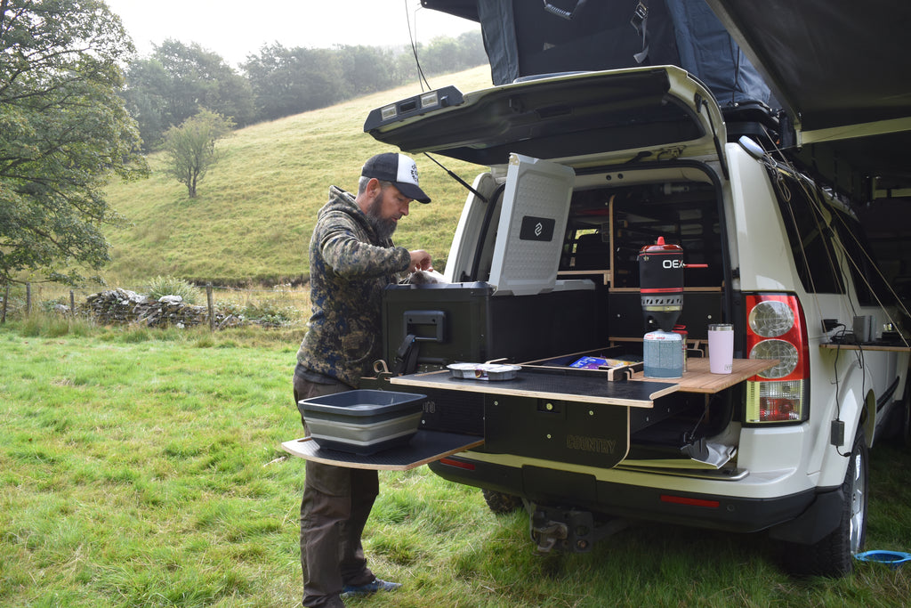Person preparing food in a Discovery 3 bed with Hexamoto Country system and camping gear in a natural setting