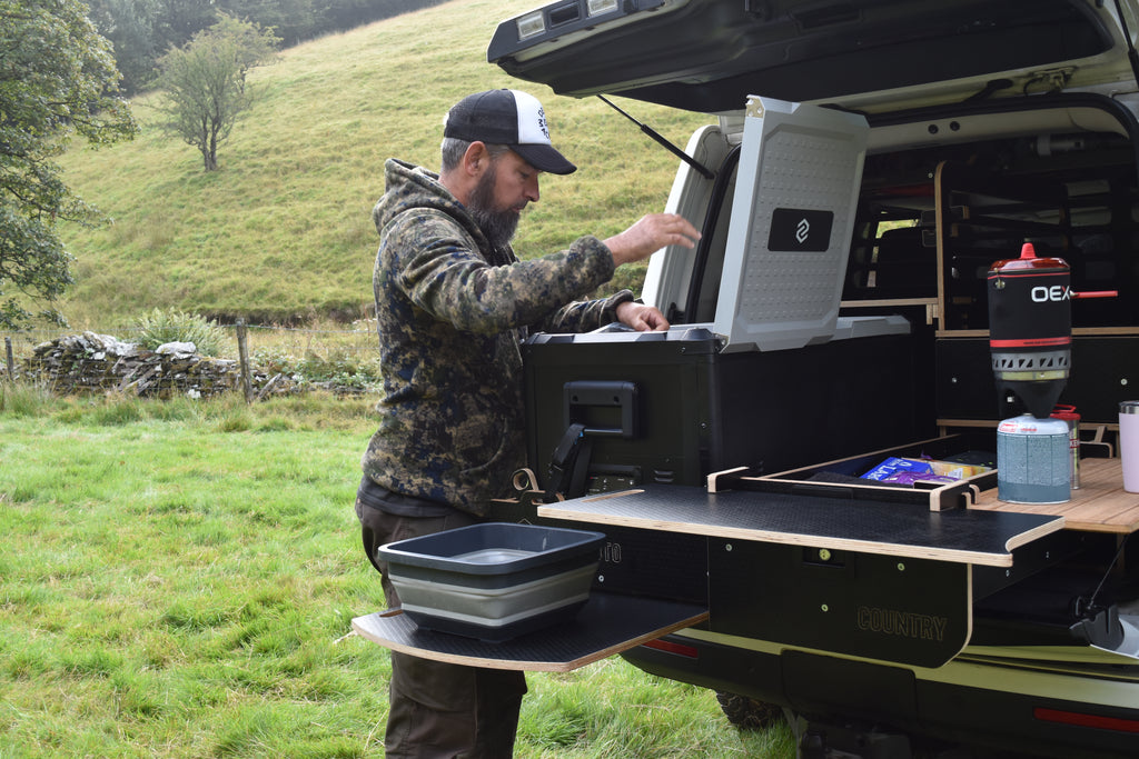 Man setting up a camping setup in the back of a vehicle with a cooler and other gear.