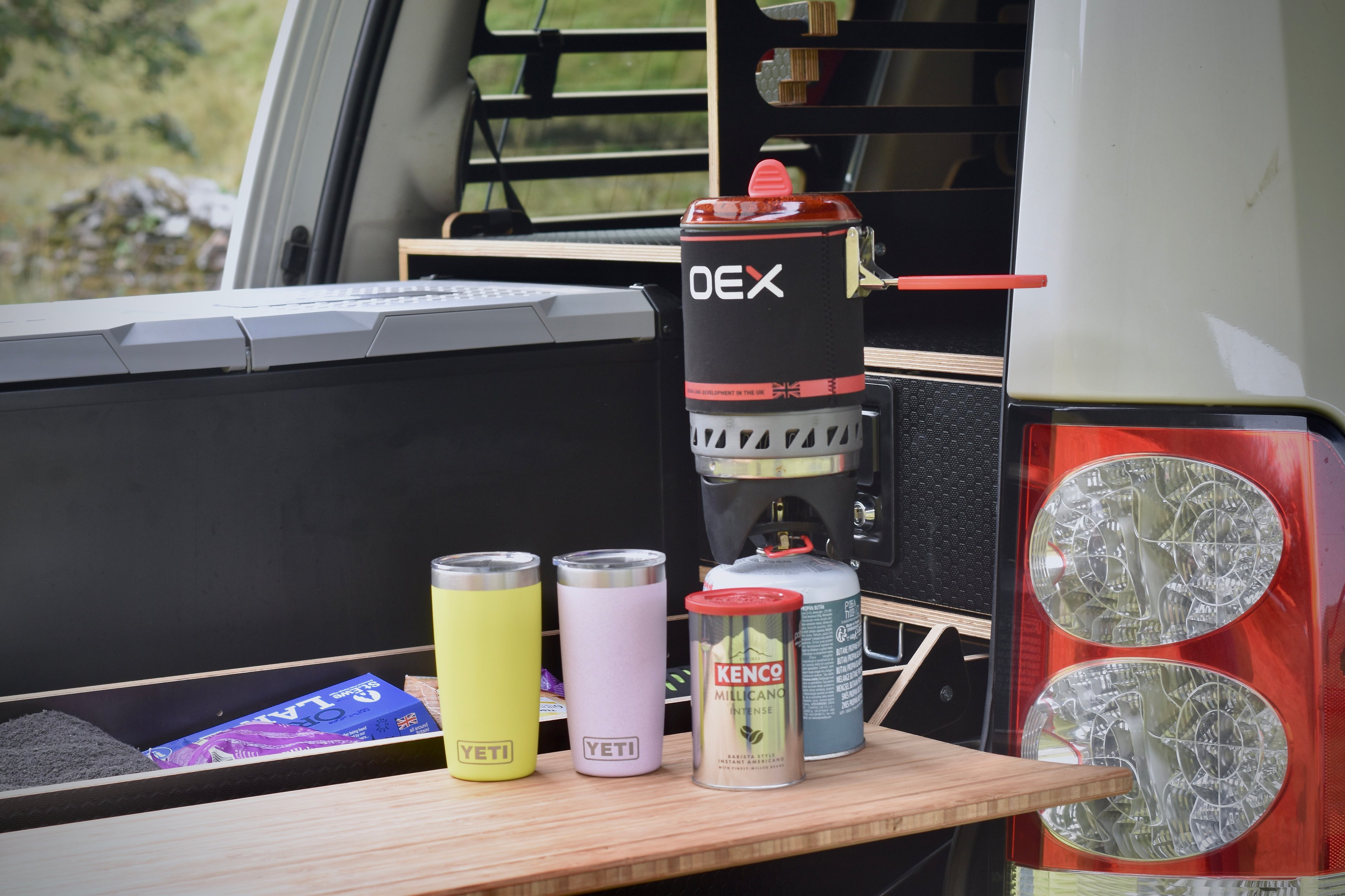 Outdoor camping setup with a portable stove, cups, and a canister of coffee on a vehicle's open rear door.