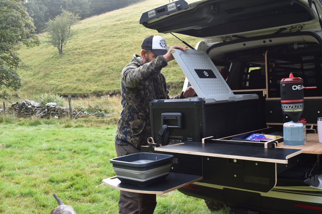 Man setting up a camping setup on a vehicle in a natural outdoor setting, opening the fridge to get food