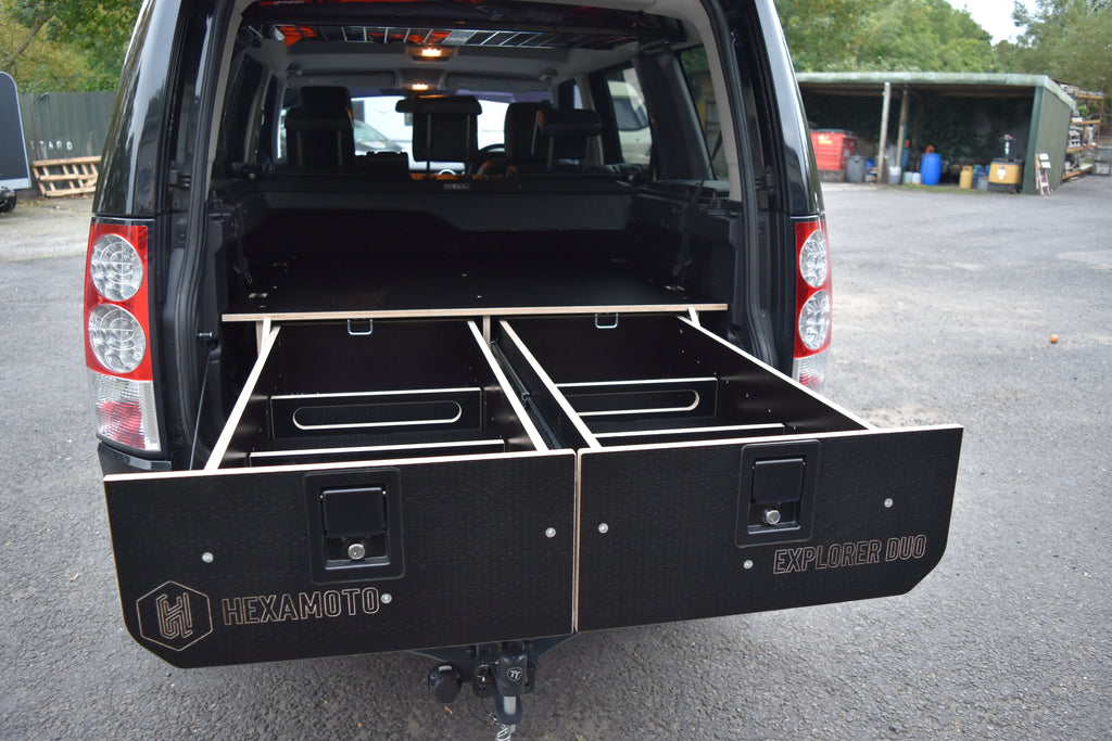 Black vehicle with open storage compartments labeled 'Hexamoto' on a paved surface.