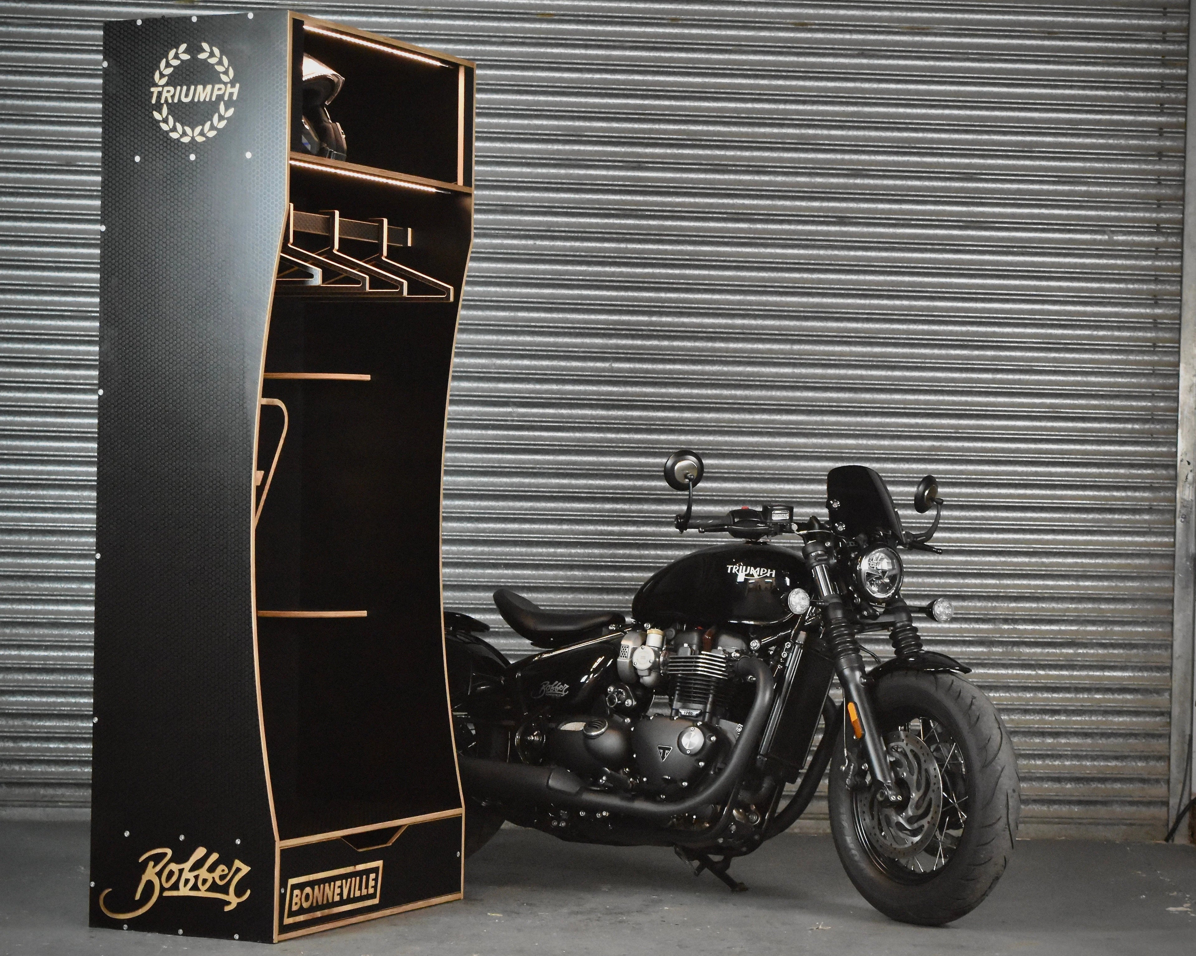 Black Triumph Bonneville Bobber motorcycle in front of a branded display stand against a metallic shutter.