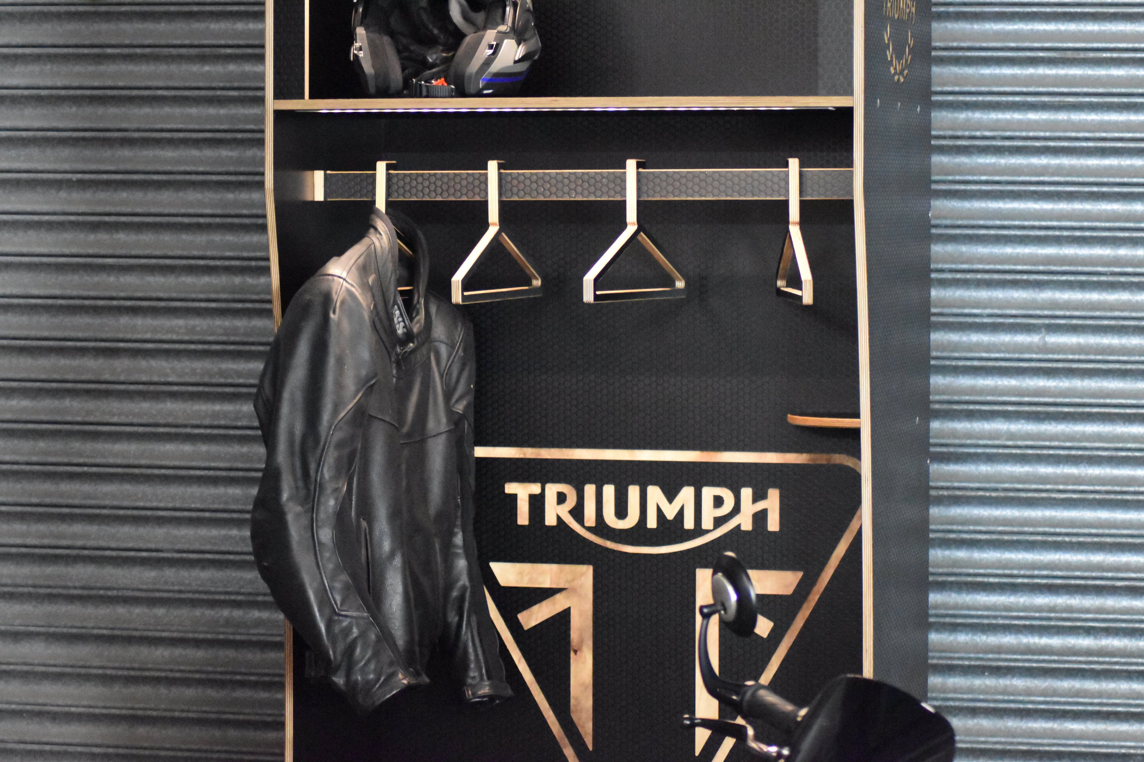 Closet with 'Triumph' branding on a black background and leather jacket hanging with helmet sat above on a shelf