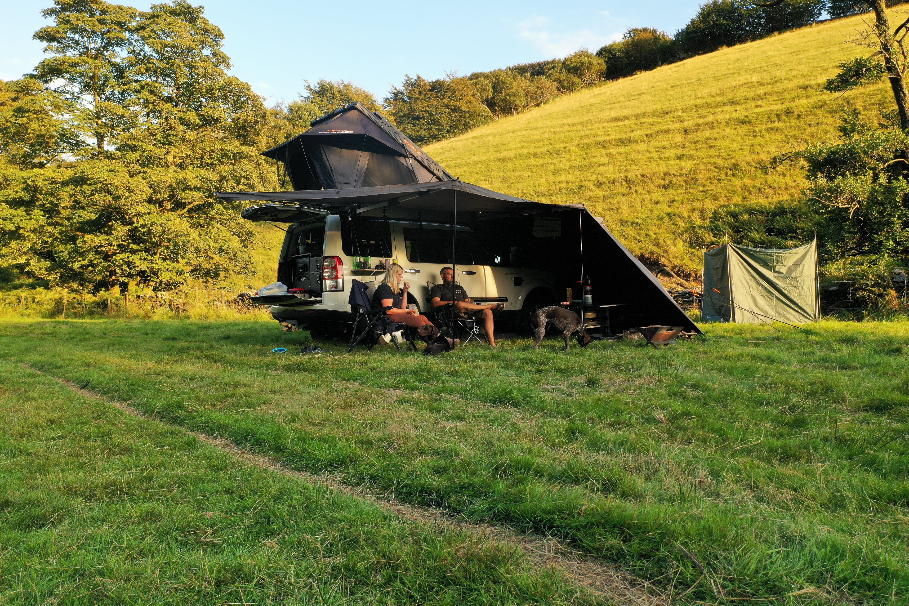 Two people sitting under a large black awning in a grassy field with trees and hills in the background.