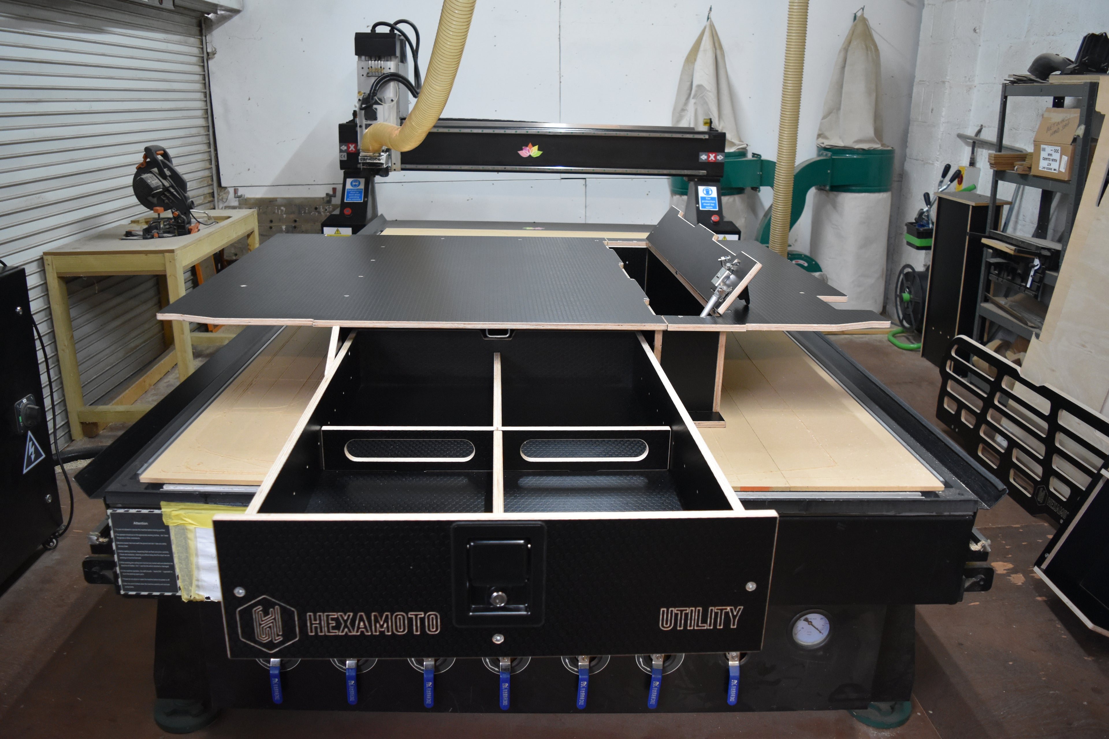 Large CNC machine in a workshop setting with various tools and equipment around and a Hexamoto Utility Storage System on top of the CNC bed with large drawer open