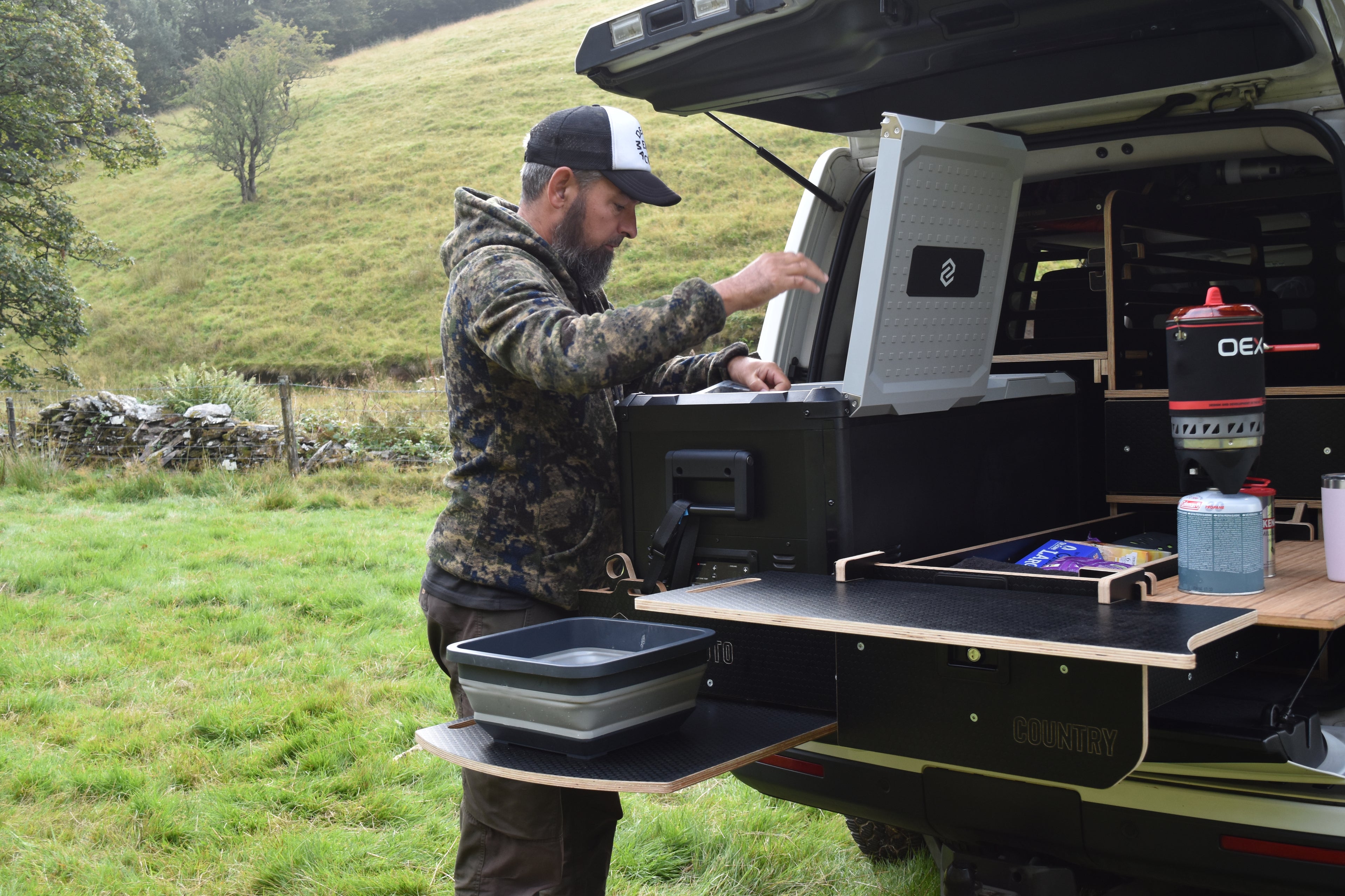 Man setting up a camping setup in the back of a vehicle with a cooler and other gear.